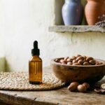Flacon compte-gouttes d'huile d'argan dorée, bol d'amandons et noix d'argan sur une natte tressée - illustration, La Savonnerie de Nyons